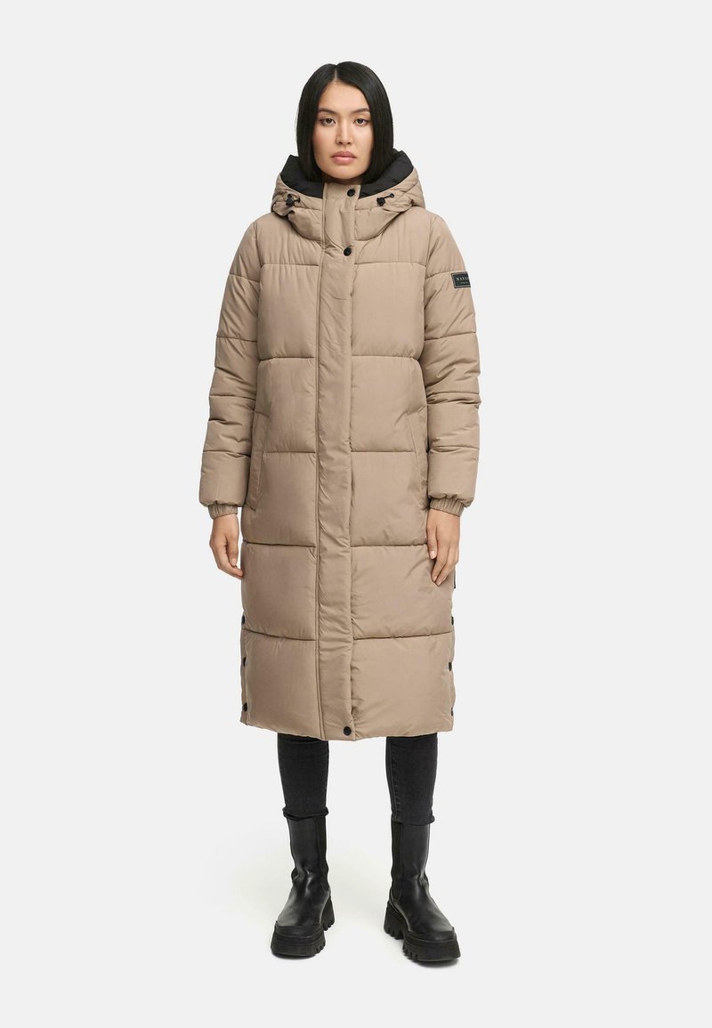 Beige lange pufferjas met een aansluitende capuchon, gestikte textuur, drukknoopsluiting en zwarte accenten. Gepaard met zwarte broeken en chunky laarzen.