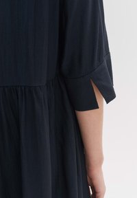 Dunkelblaues, strukturiertes Kleid mit einem gestuften Design; verfügt über Ärmel bis zum Ellbogen mit seitlichen Schlitzen. Sanfter, fließender Stoff.