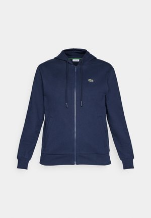 Tamsiai mėlynas zip-up gobtuvas su raišteliu, minkšta tekstūra, raukšlėtos rankogaliai ir mažas žalias logotipas kairėje krūtinės pusėje.