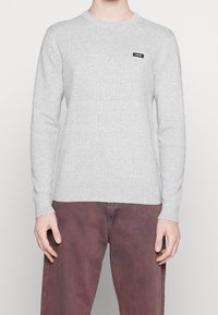 Maglione grigio a coste con maniche lunghe, caratterizzato da un collo rotondo e una piccola etichetta nera sul petto. Indossato con pantaloni color ruggine.