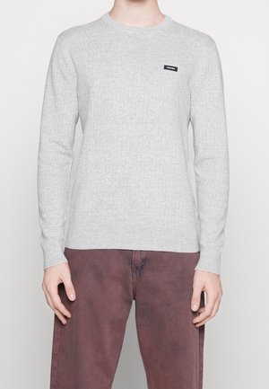 Grauer gerippter Strickpullover mit langen Ärmeln, ausgestattet mit einem Rundhalsausschnitt und einem kleinen schwarzen Logo-Tag auf der Brust. Getragen mit rostfarbigen Hosen.