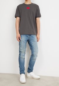 Szara koszulka z czerwoną naszywką logo, w połączeniu z jasnoniebieskimi dżinsami z przetarciami i białymi sneakersami. Tkanina wydaje się gładka, a krój jest luźny.