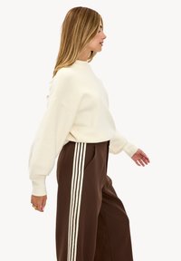 Pull crème à coupe décontractée et texture côtelée, associé à un pantalon large marron avec des rayures blanches verticales sur les côtés.