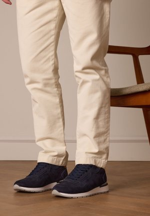 Marineblauwe suède sneakers met een gewatteerde kraag, vetersysteem en rubberen zool, gecombineerd met crème-kleurige slim-fit broek. Houten stoel zichtbaar.