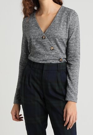 Femme portant un haut gris style cache-cœur avec trois boutons en bois et un pantalon à carreaux foncé taille haute, debout devant un fond uni.