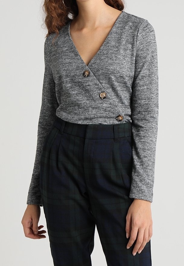 Femme portant un haut gris style cache-cœur avec trois boutons en bois et un pantalon à carreaux foncé taille haute, debout devant un fond uni.