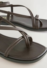 Tmavohnedé ploché sandále s tenkými koženými remienkami prekríženými do tvaru X a štvorcovým dizajnom špičky na bielom pozadí.