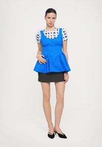 Blue peplum top with gathered details, worn over a black and white checkered shirt. Paired with a patterned black skirt and black shoes.