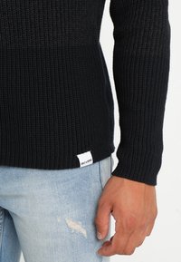 Jersey negro de punto con textura acanalada y mangas largas, con una etiqueta lateral. Combinado con un denim desgastado de color azul claro.