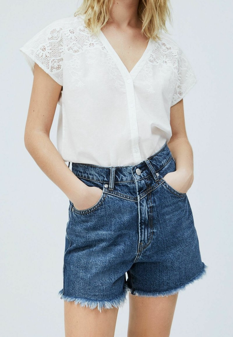 Bílá blůza s krátkým rukávem, zdobená vyšívanými detaily a s výstřihem do V, v kombinaci s modrými denimovými šortkami s vysokým pasem a roztrhanými lemy.