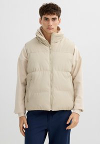 Chaleco acolchado beige con cuello alto, cierre delantero de cremallera y dos bolsillos laterales, llevado sobre una sudadera de color claro. Textura suave, ajuste informal.