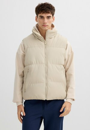 Chaleco acolchado beige con cuello alto, cierre delantero de cremallera y dos bolsillos laterales, llevado sobre una sudadera de color claro. Textura suave, ajuste informal.