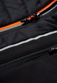 Schwarzer gesteppter Stoff mit orangefarbenen Reißverschlussakzenten und reflektierenden Streifen. Verfügt über mehrere Reißverschlusstaschen und eine strukturierte Oberfläche.