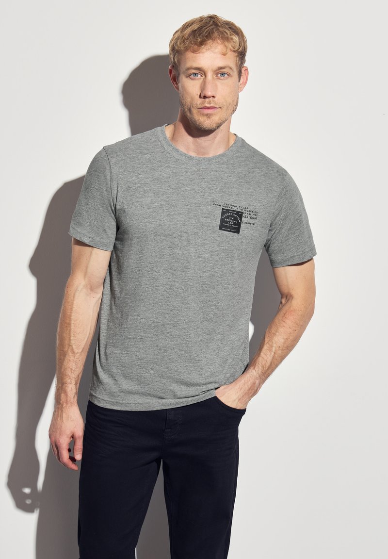 Šedé bavlněné tričko s krátkým rukávem, s černým čtvercovým designem a textem na levé hrudi. Model má na sobě námořnické kalhoty, ruce v kapsách.