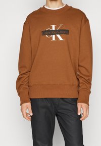 Sudadera marrón con un logotipo gráfico, cuello y puños acanalados. Combinada con pantalones oscuros, que presentan un ajuste relajado y una textura suave.