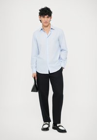 Jasnoniebieska koszula w paski na guziki z długimi rękawami, czarne dopasowane spodnie, czarne buty z białymi akcentami, trzyma czarną kopertówkę.