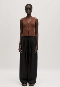 Brauner gestrickter ärmelloser Oberteil mit V-Ausschnitt und Knopfverschluss, kombiniert mit schwarzen weiten Hosen und schwarzen Sandalen. Glatte Textur.