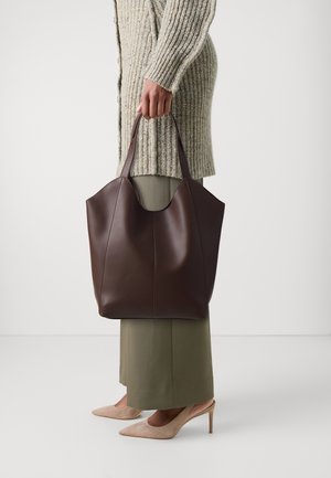 Femme tenant un grand sac cabas en cuir brun foncé, vêtue d'un pull en maille beige, d'une jupe olive et de talons pointus beiges, sur fond uni.