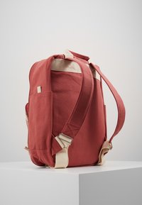 Sarkans audekla mugursoma ar bēšiem siksnām. Iekļauj sānu kabatu, polsterētu aizmuguri un regulējamas siksnas pielāgošanai. Tekstūrēta virsma un minimālistisks dizains.