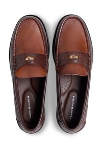 Καφέ δερμάτινα μοκασίνια με λεία επιφάνεια, που διαθέτουν διακοσμητικό λουράκι και χρυσές μεταλλικές λεπτομέρειες. Υφασμένο πάτο με τρύπες για μεγαλύτερη άνεση.