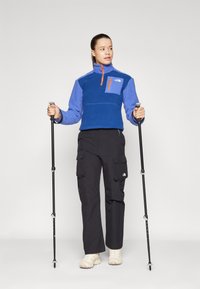 Plava flis jakna s visokim ovratnikom i zatvorenim džepom, uparenim s crnim cargo hlačama i planinarskim štapovima; teksturirani materijal i sportski dizajn.