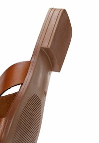 Brun sandal med en texturerad gummisula, med en bred remdesign och platt häl. Sulan har spår för grepp och stabilitet.