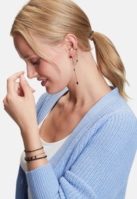 Orecchini a catena neri con accenti di perle, indossati con un cardigan lavorato a maglia blu chiaro. Il bracciale presenta un design simile in nero, abbinato a una maglietta bianca.