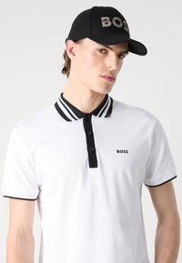 White polo shirt with black stripes on the collar and black buttons. "BOSS" logo in black on the left chest. Black cap with "BOSS" logo.