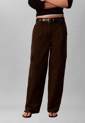 Kobieta w czarnym krótkim swetrze, ciemnobrązowych szerokich spodniach typu cargo z paskiem i czarnych sandałach z odkrytymi palcami, stoi z założonymi rękami.