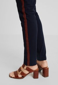 Jambes portant un pantalon bleu marine avec des bandes latérales rouges et des sandales marron à talons avec deux brides à boucles, debout sur un sol gris clair.