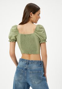 Olivengrünes, kurzes Oberteil mit Puffärmeln und gerafftem Rückendetail, kombiniert mit hochgeschnittenen blauen Jeans, die einen strukturierten Stoff zeigen.