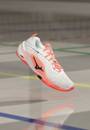 Chaussure de sport blanche et corail avec un logo noir flottant au-dessus d'un sol de terrain intérieur marqué de lignes colorées.