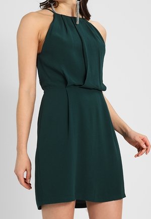 Femme portant une robe sans manches, vert foncé, longueur genou, avec un col froncé et de fines bretelles, debout devant un fond uni.