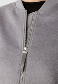 Nahaufnahme eines silbernen Reißverschlusses auf einer grauen Wolljacke, die über einem schwarzen Oberteil getragen wird, mit einem grauen Stoff-Zipp-Anhänger, der an dem Reißverschluss-Schieber befestigt ist.