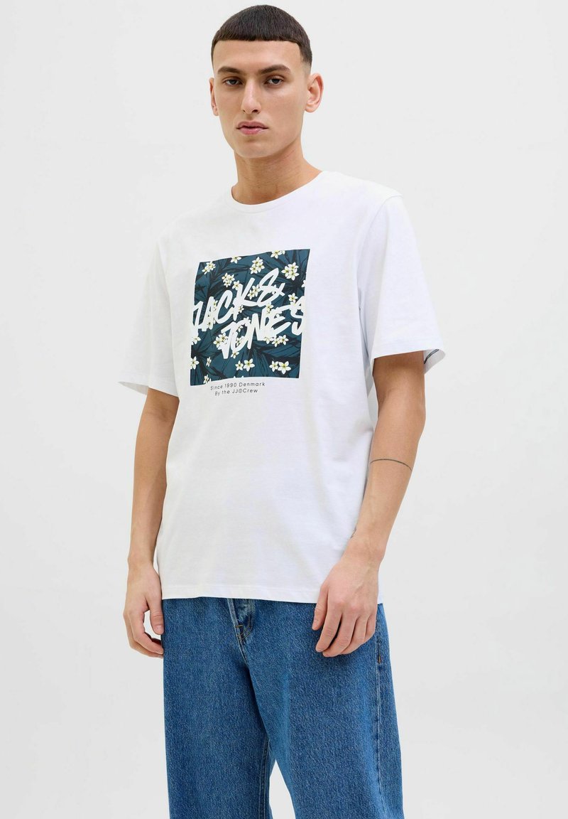 Bílá bavlněná tričko s námětem čtvercového grafického vzoru květin a textem "JACK & JONES" tučně, nošené s volnými modrými džíny.