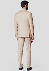 Beżowy garnitur o dopasowanym kroju, z jednym rozcięciem, dopasowanymi spodniami i czarnymi butami, widziany z tyłu. Gładka, teksturowana tkanina.