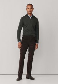 Dunkelgrüner Zip-Pullover aus weichem Material, kombiniert mit braunen Cordhosen. Das Modell trägt schwarze Schuhe und ein hellblaues Hemd darunter.