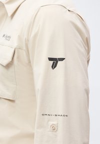 Κοντινό πλάνο μανσετών jacket σε μπεζ χρώμα με μαύρο γεωμετρικό λογότυπο και κείμενο «OMNI-SHADE» πάνω από ένα μικρό κουμπί.