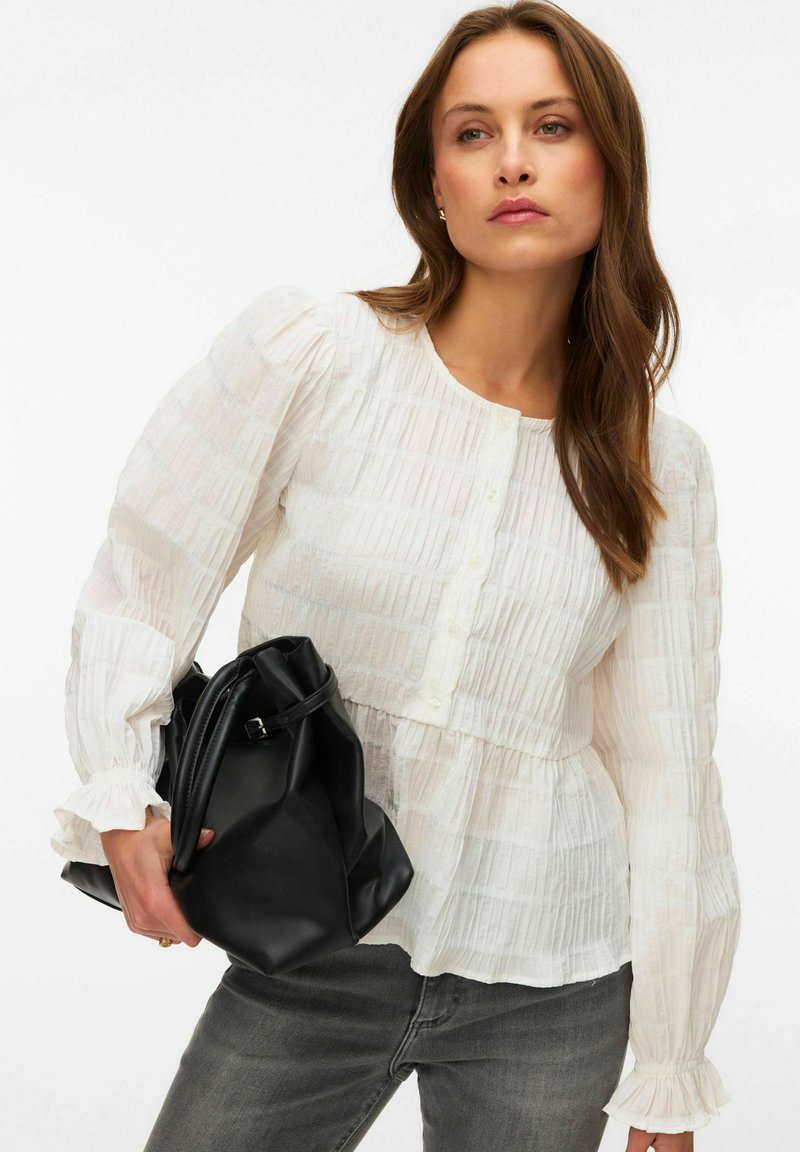 Femme portant un chemisier blanc texturé et un jean gris, tenant un grand sac à main en cuir noir sur un fond uni.