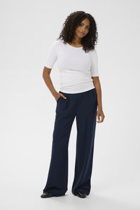 Sieviete ar cirtainiem matiem, tērpusies baltā pieguļošā īsroku kreklā, vaļīgās tumši zilās biksēs un melnās dzīvokļveida kurpēs, stāv ar vienu roku kabatā.