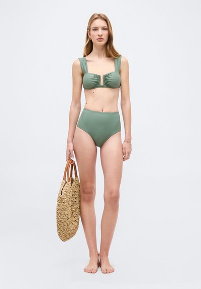 Γυναίκα που φοράει πράσινο μαγιό με ψηλή μέση, κρατώντας ένα υφαντό ψάθινο τσάντα με καφέ λαβές, όρθια ξυπόλητη σε ένα απλό ανοιχτό γκρι φόντο.