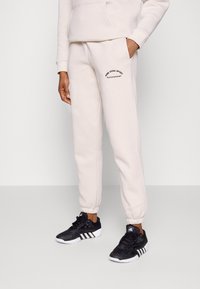 Beige sweatpants med elastiska muddar, prydda med en svart logotyp på vänster lår. Åtföljd av svarta sportskor med vita sulor.