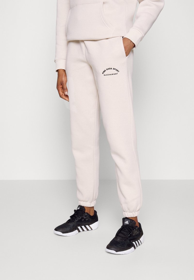 Beige sweatpants med elastiska muddar, prydda med en svart logotyp på vänster lår. Åtföljd av svarta sportskor med vita sulor.