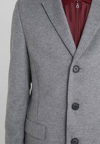 Abrigo de lana gris con un diseño ajustado, que presenta un cuello con muescas, dos bolsillos frontales y botones negros. El forro interior es burdeos con rayas.