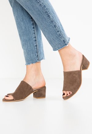 Pieds portant des mules à talons blocs ouverts en daim marron avec un jean bleu longueur cheville sur fond blanc.