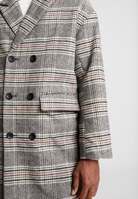 Double-breasted wool coat featuring a gray and white houndstooth pattern with red accents, black buttons, and a side pocket.