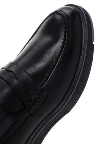 Zapato de cuero negro con un acabado suave, que presenta una correa en la parte delantera, suela de goma texturizada y detalles de costura sutiles a lo largo de los bordes.
