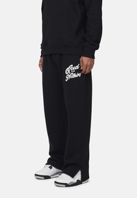 Zwarte katoen hoodie met een voorzak en bijpassende zwarte trainingsbroek met witte tekst "Good For Nothing", gecombineerd met witte sneakers.