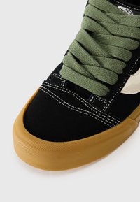 Sneaker nera in suede con lacci verdi, logo bianco e suola in gomma testurizzata color corda. Presenta cuciture bianche lungo la parte superiore.
