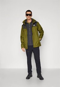 Zelena nepremočljiva jakna s črnimi poudarki, oblečena čez sivo flis jakno z zadrgo, kombinirana s temnimi hlačami in črnimi čevlji. Model stoji sproščeno.